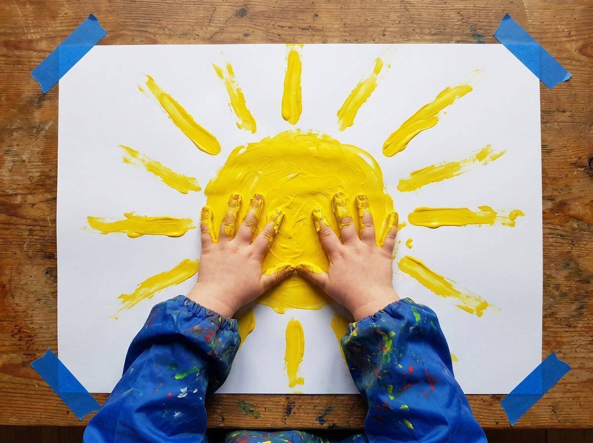 Manos pequeñas pintando con dedos amarillos los rayos del sol sobre papel — actividad Los Mundos de Noa