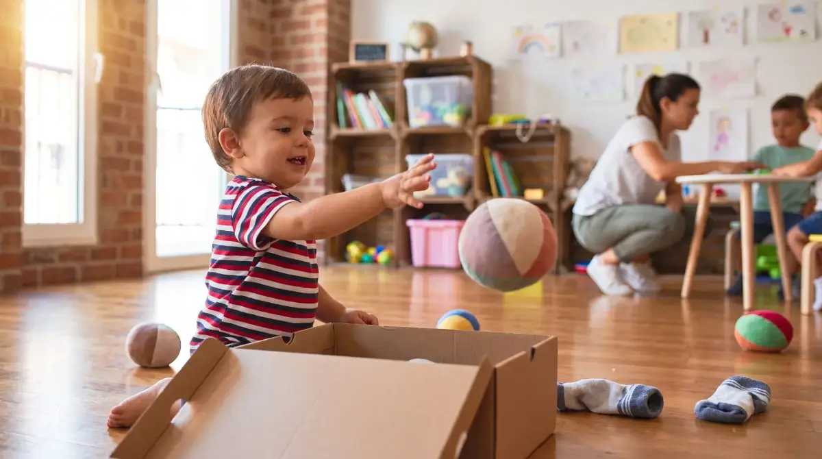 Niño de año y medio lanzando una pelota de tela dentro de una cesta de cartón en un aula infantil — cesta de lanzar