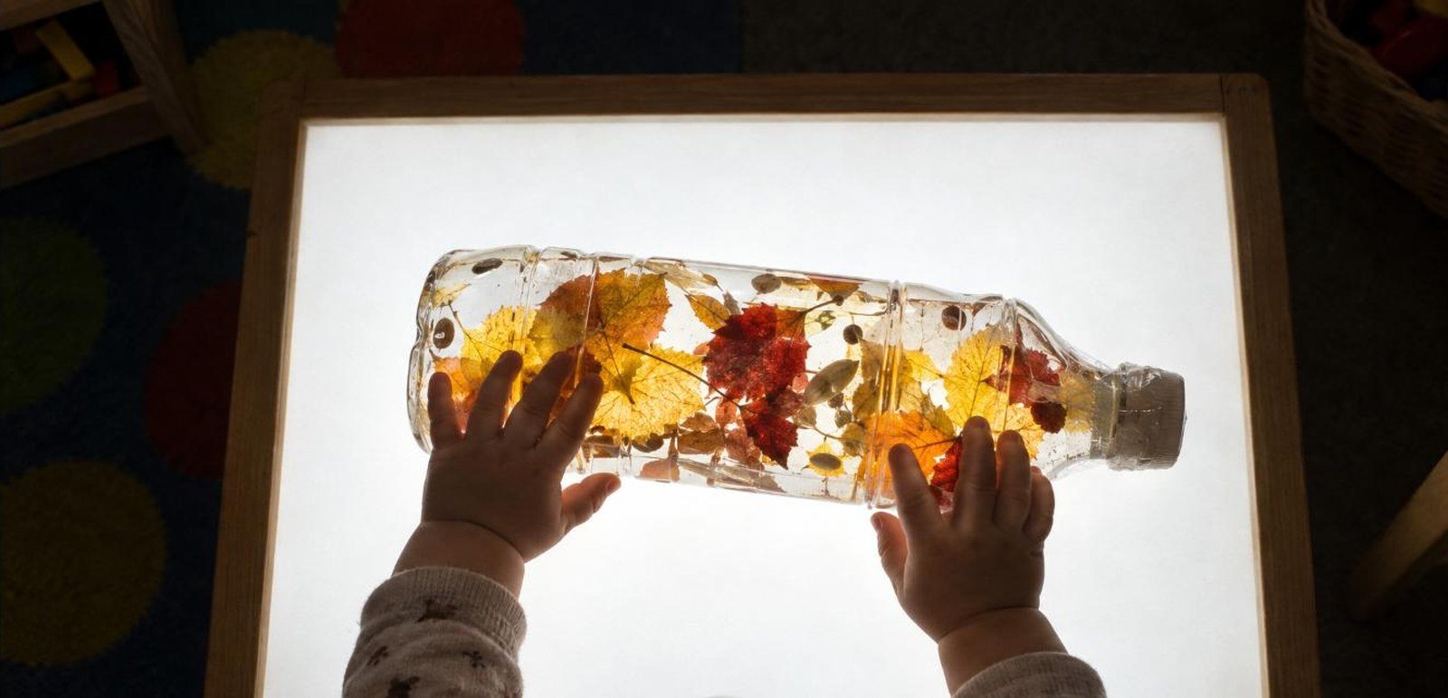 Bebé de 12 meses empujando con las dos manos una botella sensorial con hojas de otoño sobre la mesa de luz — botella sensorial de otoño