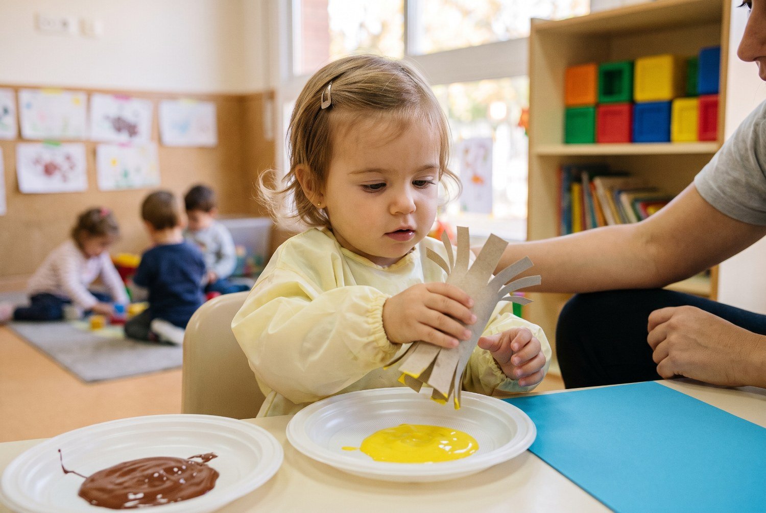 Niña de 1 año tocando las tiras abiertas de un rollo de cartón junto a un plato de témpera amarilla