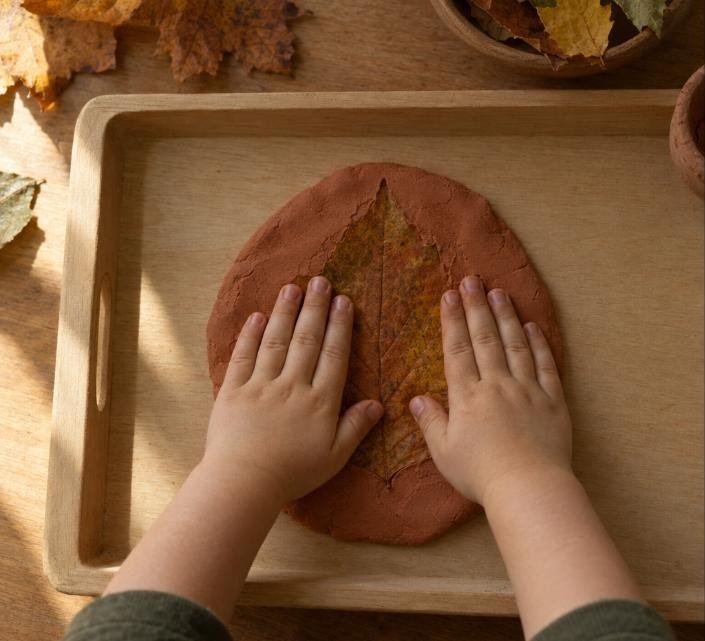 Manos infantiles presionando una hoja seca de otoño contra un disco de color terracota
