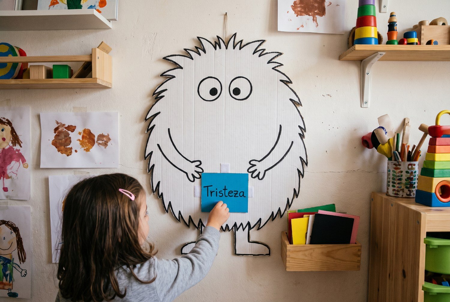 Niña de 4 años pegando con velcro una tarjeta azul en la tripa de un monstruo de cartulina colgado en la pared del aula