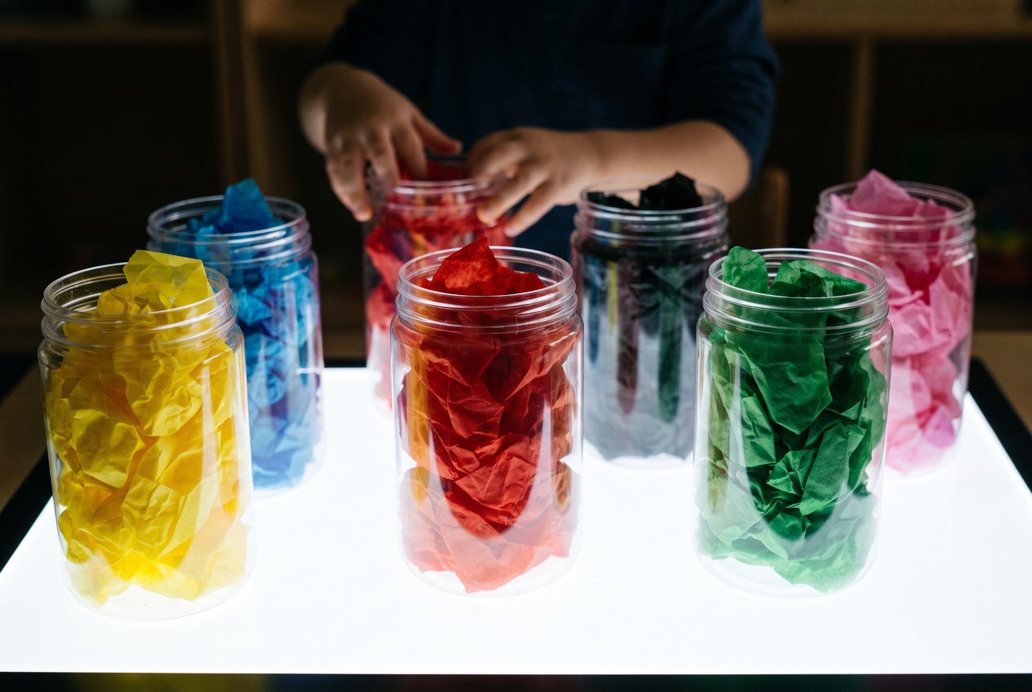 Seis botes translúcidos llenos de papeles de colores iluminados desde atrás en una mesa de luz