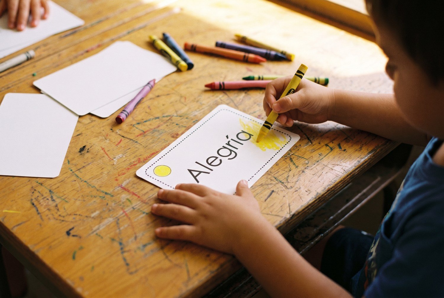 Manos de un niño de 3 años coloreando de amarillo una tarjeta con la palabra alegría junto al cuento abierto