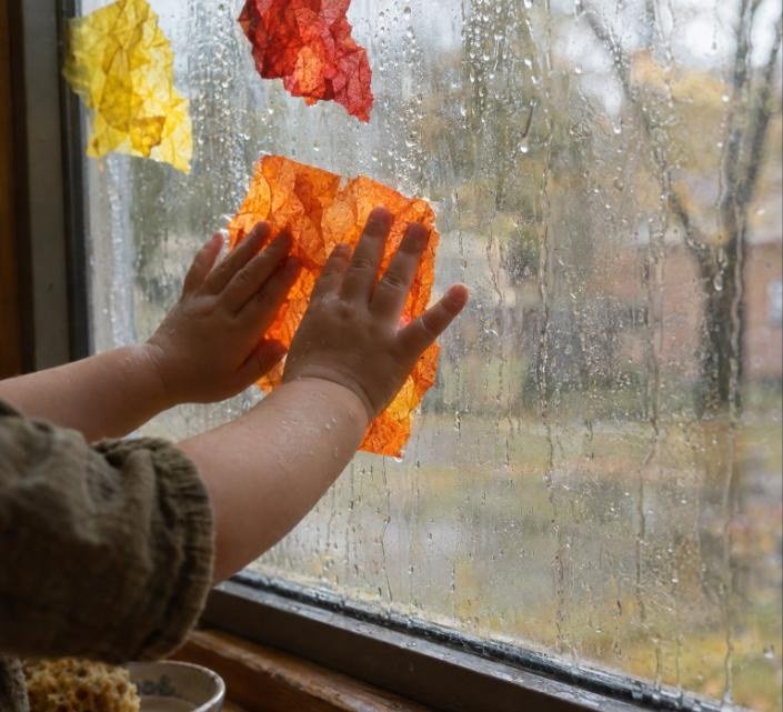 Bebé pegando un trozo de papel de seda naranja sobre el cristal mojado de la ventana