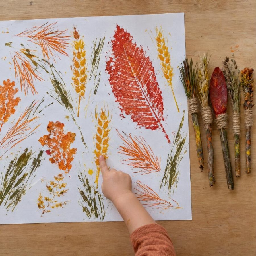 Papel lleno de huellas de otoño y cinco pinceles de naturaleza manchados de pintura; el niño señala una huella