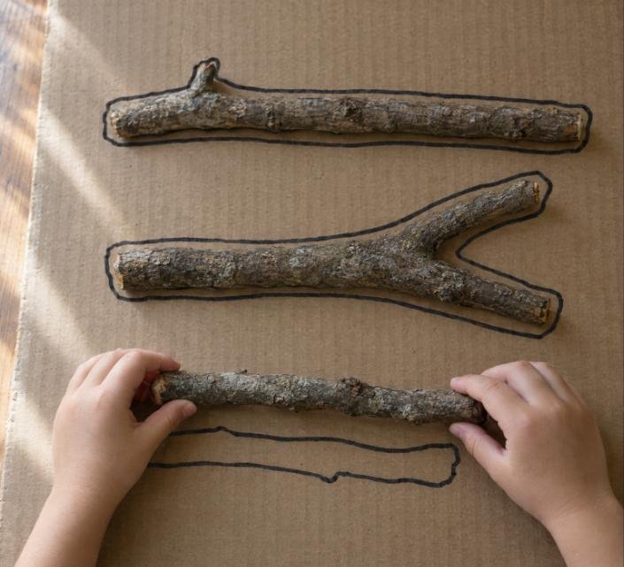 Actividad con ramas: manos de un niño colocando un palo sobre su silueta dibujada
